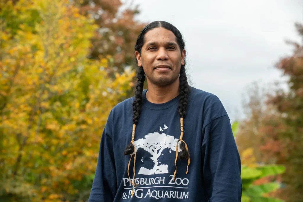 sunbear standing outside at the zoo. he is wearing a zoo sweatshirt and his hair is in braids.