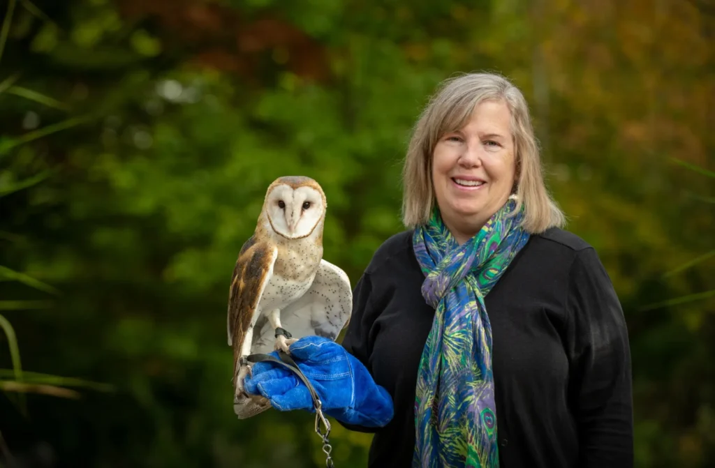 margie standing with an owl perched on her gloved hand. margie has light colored hair and is wearing a peacock patterned scarf.