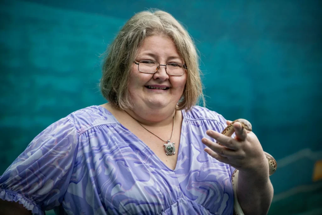 beth holding a small amassador animal snake in her hand. beth has short light colored hair and is wearing glasses, a swirly purple shirt, and green turtle necklace.