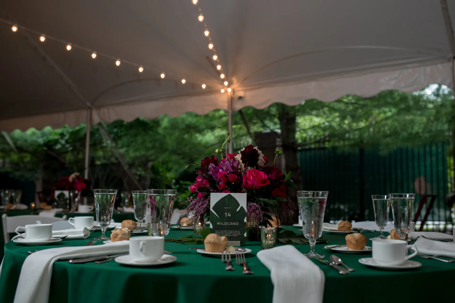 a round table set up in the garden tent. the table is set for 10 guests. the tablecloth is dark green, and there are white napkins, coffee cups, silverware, water glasses, and rolls with butter on the table. a centerpiece with a tropical plant leaf and flowers is in the middle of the table. the tent is overhead with string lights.