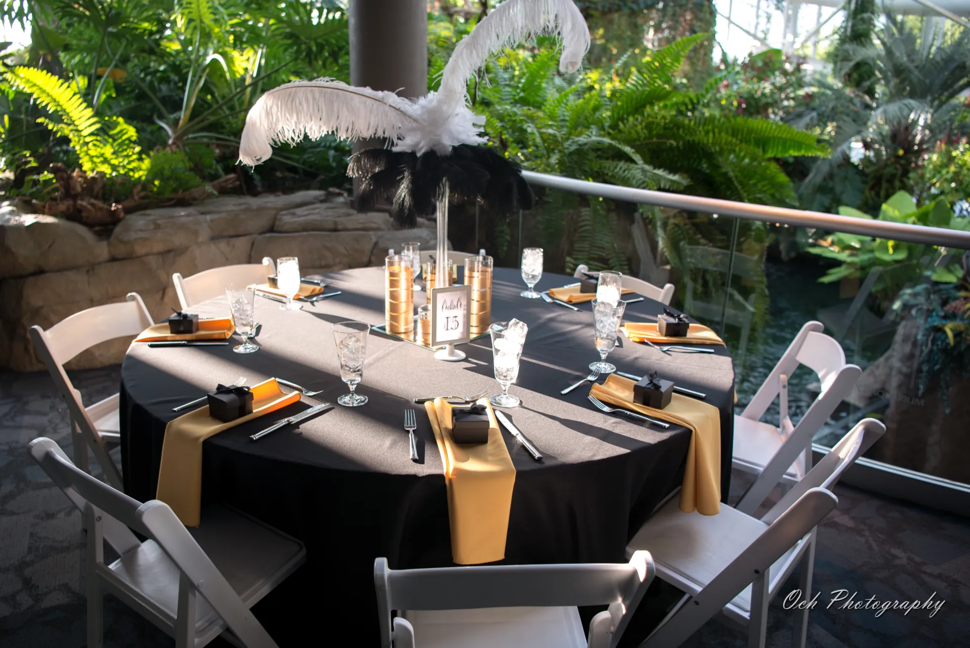 a table set for seven guests in the upper aquarium. the linens are black with gold napkins and small black giftboxes. there is a large ostrich feather as the table centerpiece.