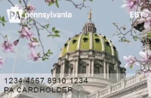 pennsylvania ebt card. in the top left, the card has the letters PA in a keystone shape next to the word Pennsylvania. it says EBT in the top right. across the bottom are cardholder numbers and a cardholder name. a picture of the capitol building in harrisburg with cherry blossoms is in the background.