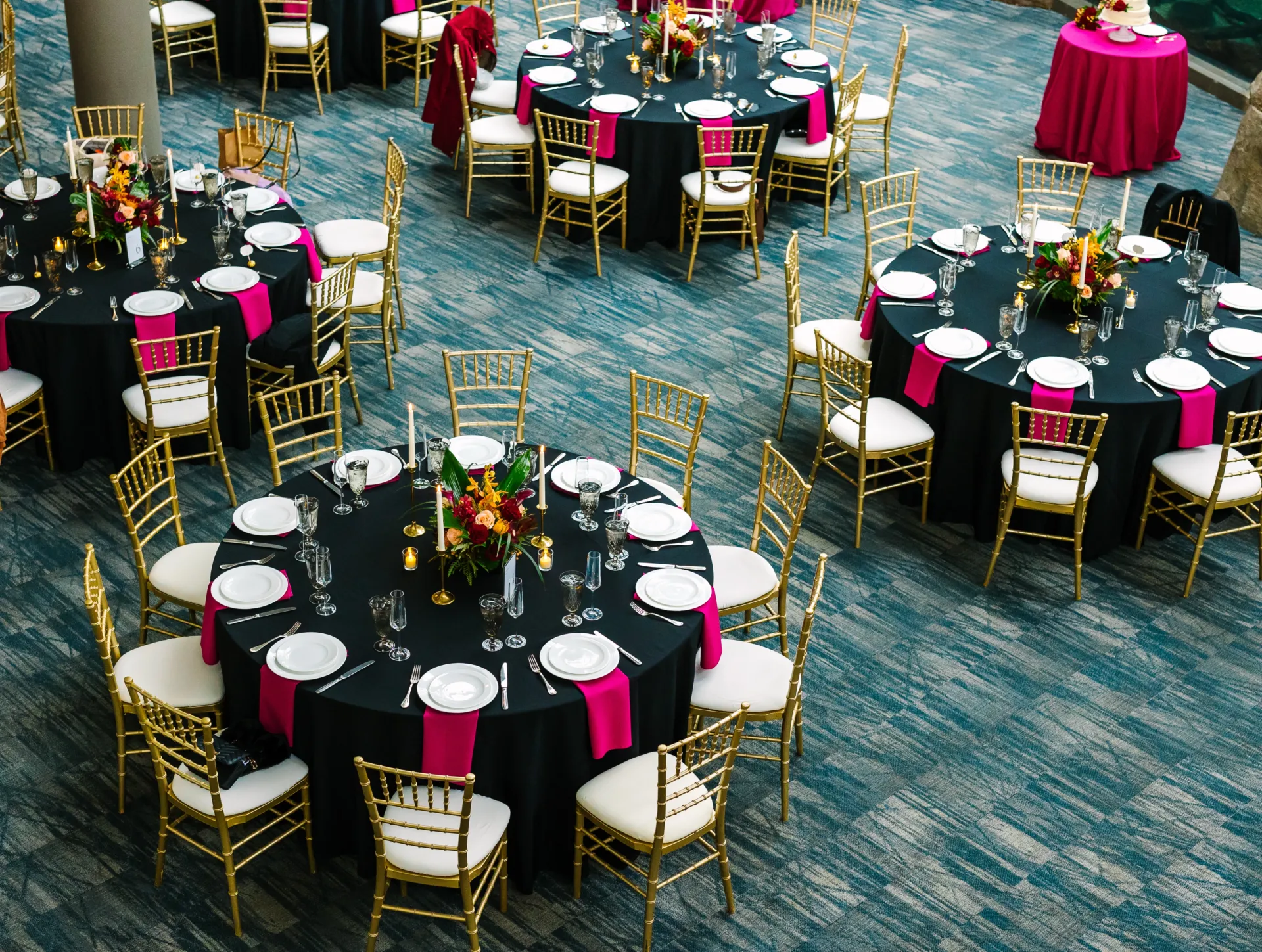 a selection of four tables set up on the lower floor of the aquarium. the tables are set for ten guests, with a dark blue table cloth and hot pink napkins that pop brightly. tropical flowers are in the center of the tables.